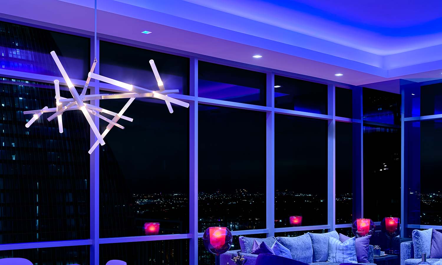 Modern living room at night with floor-to-ceiling windows, city views, blue accent lighting, and a contemporary chandelier.