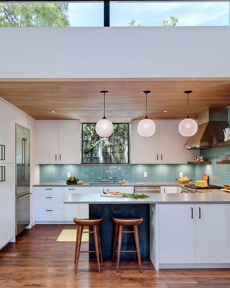 Bright contemporary kitchen with white cabinets, teal tile backsplash, wood floors and ceiling, an island with two wooden stools, and three globe pendant lights hanging beneath a high clerestory window.