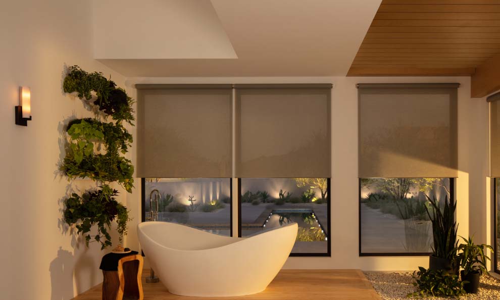 Spa-like bathroom with a sculptural white freestanding tub in front of three large windows covered by beige roller shades, a vertical wall garden on the left, potted plants on the right, and a view of an illuminated outdoor pool and landscaping at dusk.