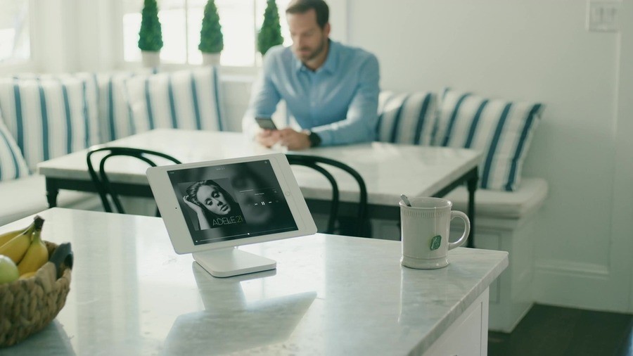 Man in dining room with tablet displaying Adele's music and a coffee mug on the kitchen island next to him. 