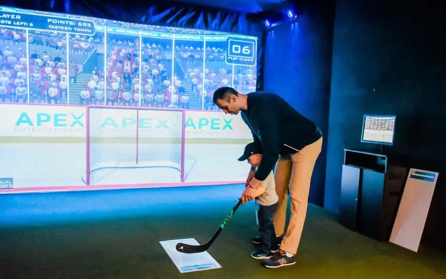 A man and a small child playing with a hockey stick in front of a sports simulator screen. 