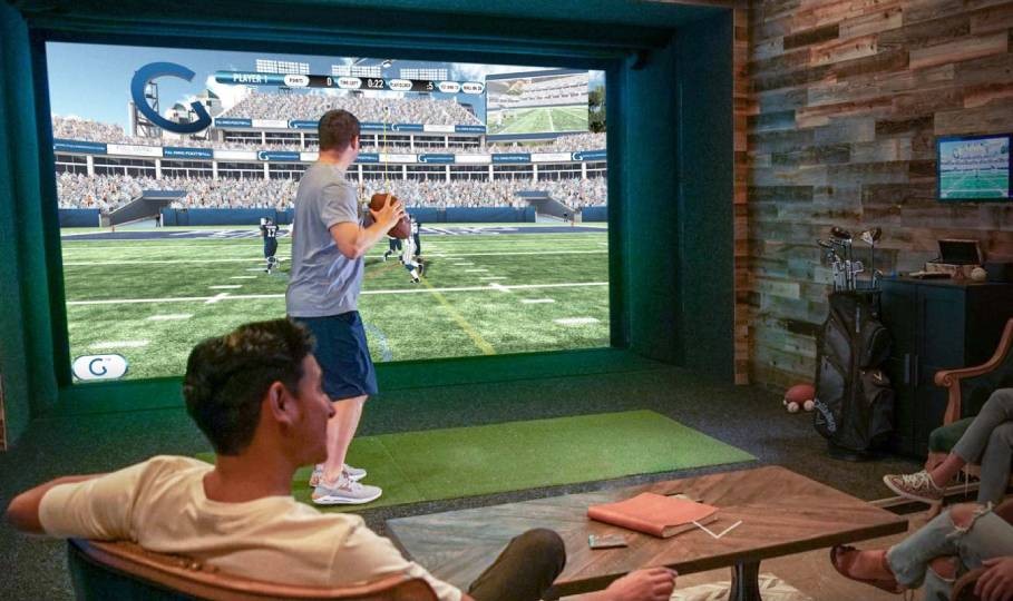 One person is throwing a virtual football at the large projected screen displaying a football field, while others watch comfortably from seating areas within the rustic, wood-paneled room. 