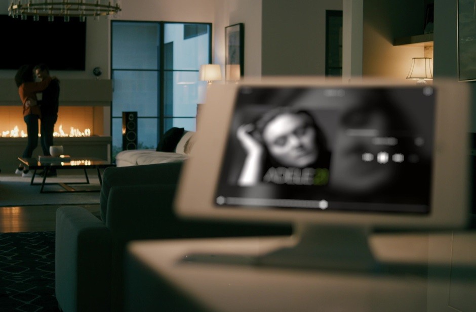A modern living room featuring a Savant touch screen displaying a music playlist with a couple embracing near a fireplace in the background.