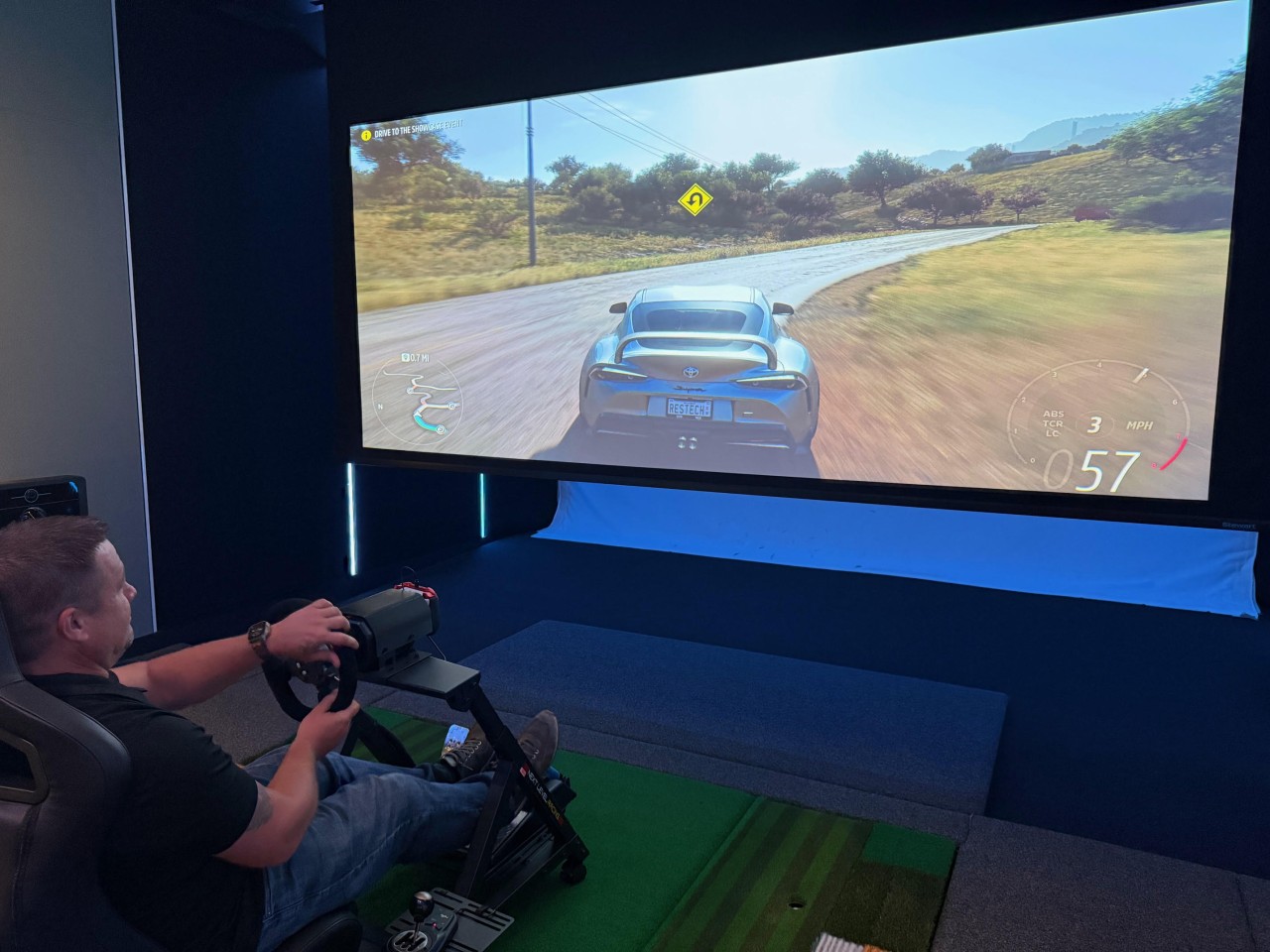 Person sitting in GTR racing seat and using a hybrid sport simulator. A car is shown on the screen.