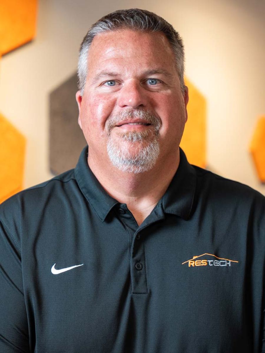 A man with short gray hair and a goatee wearing a black polo shirt with a ResTech logo and a Nike swoosh, posing for a professional headshot against a background with orange geometric wall panels.