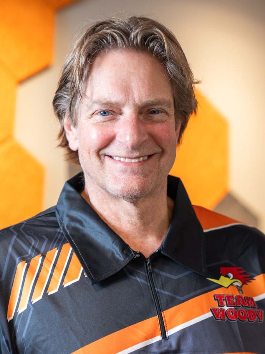 Smiling man with medium-length light brown hair wearing a black and orange “TEAM WOODY” polo shirt, standing indoors against a blurred background with orange geometric wall panels.