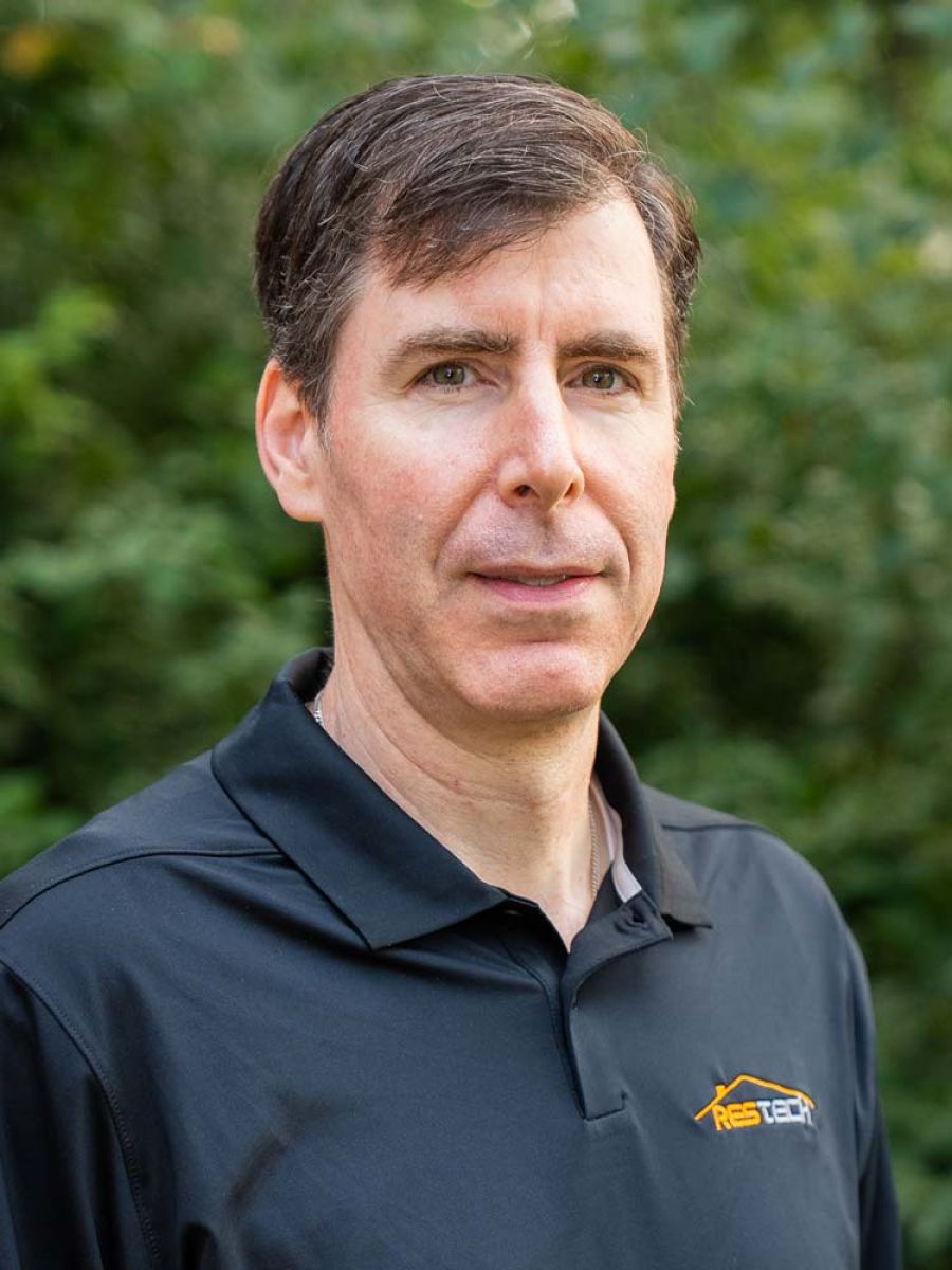 Man with short brown hair wearing a black polo shirt with an orange “ResTech” logo, standing outdoors in front of blurred green foliage.