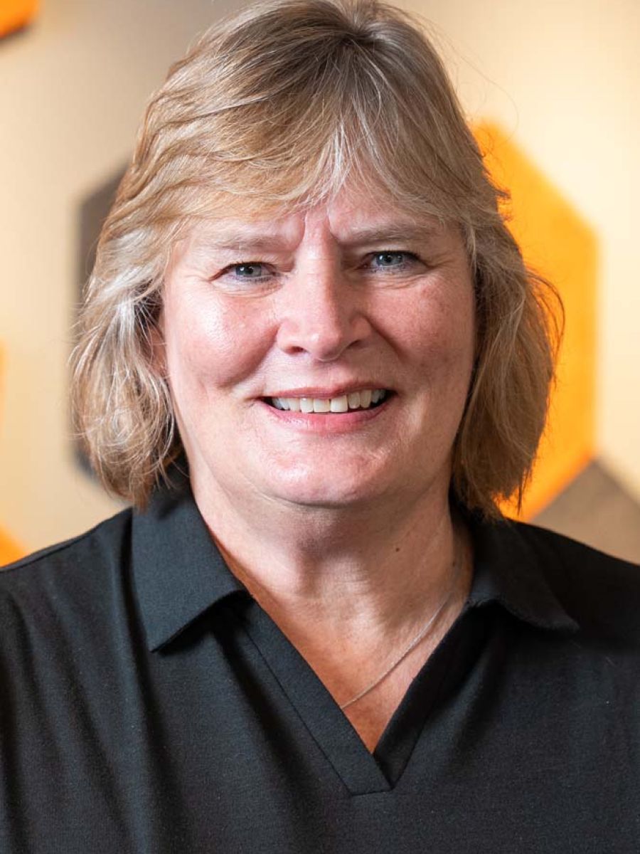 Smiling woman with short blond hair wearing a black collared shirt, standing indoors in front of a blurred background with orange geometric wall panels.