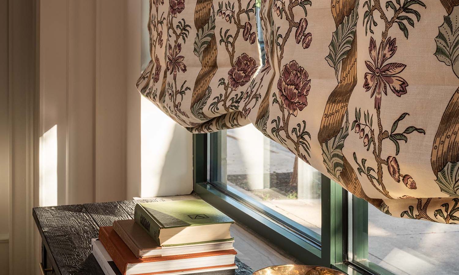 Close-up of a patterned Roman shade partially lowered over a window, with sunlight illuminating a stack of books on a nearby table.