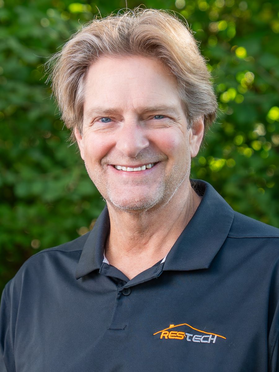 Smiling man with medium-length light brown hair wearing a black and orange “TEAM WOODY” polo shirt, standing indoors against a blurred background with orange geometric wall panels.